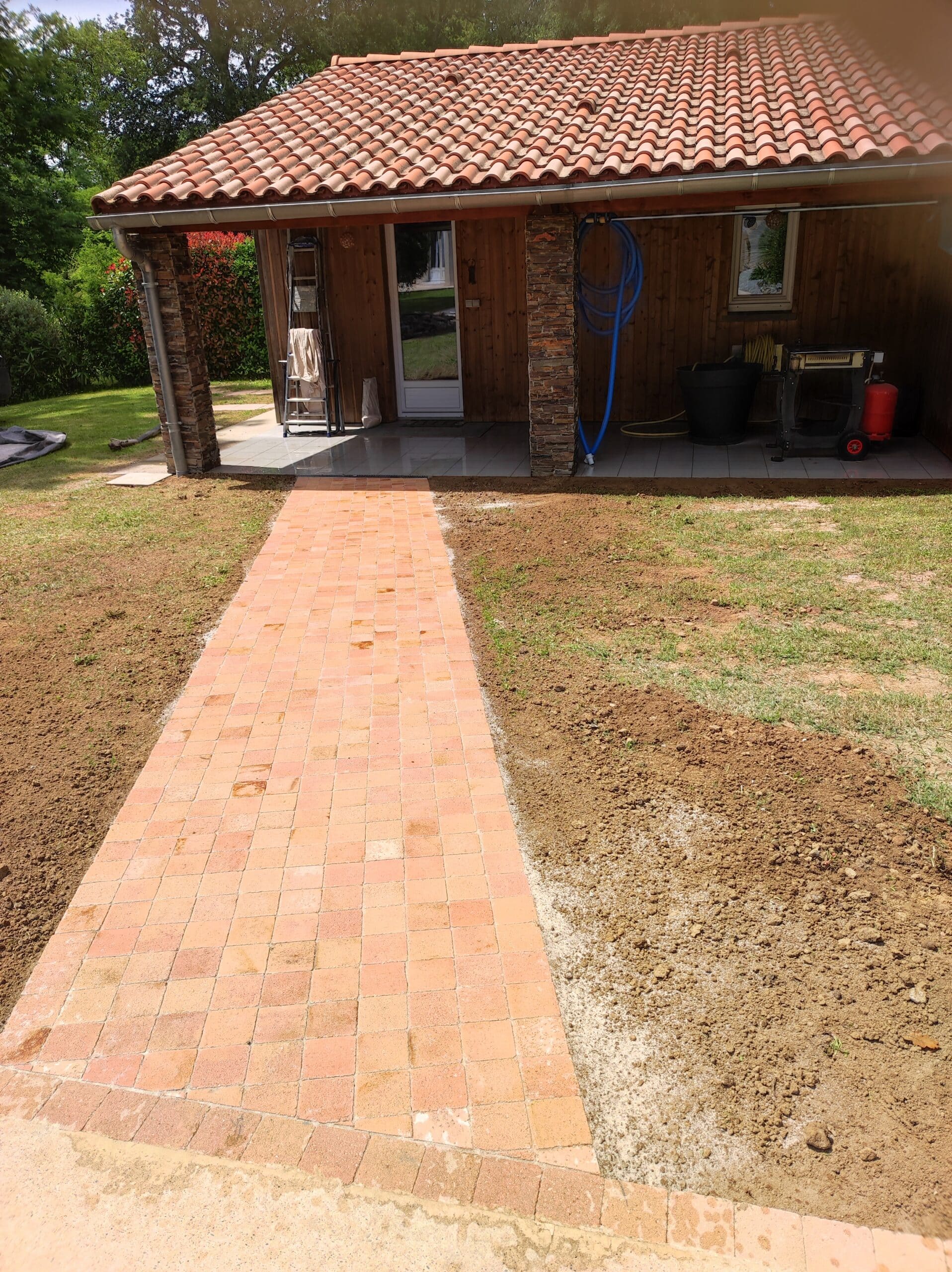 Allée de pavés en terre cuite menant à une maison avec toit de tuiles rouges et piliers en pierre naturelle.