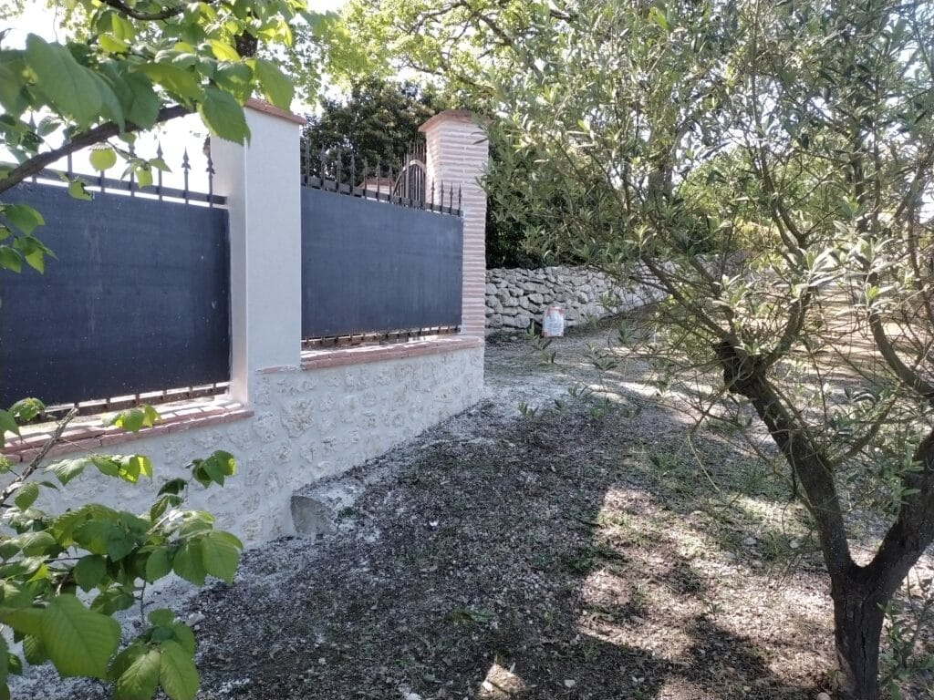 Clôture de jardin en fer forgé et muret en pierre naturelle Clôture élégante en pierre, crépi blanc et fer forgé noir bordée par un olivier et des feuillages dans un jardin ensoleillé.