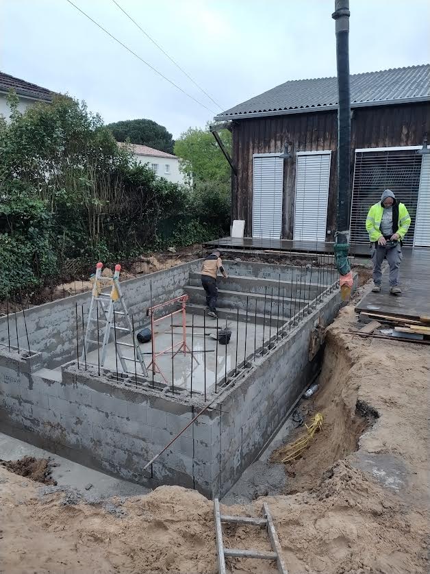 Chantier de construction d'une piscine en blocs béton avec ouvriers devant une maison en bois sombre.